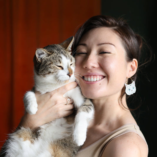 Smiling woman wearing hand-painted Persian white cat hoop earrings from Le Chic Miami while holding a tabby cat.