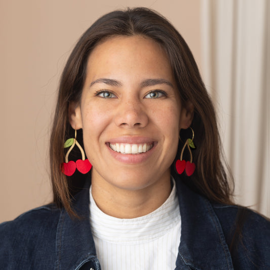 Woman wearing cherry-shaped hand-painted wooden earrings by Le Chic Miami, smiling in denim jacket.
