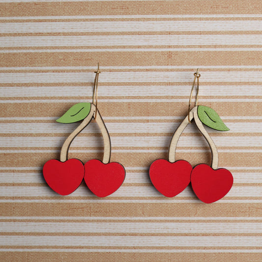 Pair of handmade wooden cherry earrings with bright red heart-shaped cherries and green leaves, displayed against a beige and white striped background, perfect for adding a playful, vibrant touch to any outfit.