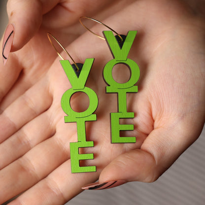 A pair of bright green novelty hoop earrings shaped like the word 'VOTE', held in a person's hands against a soft-focus gray background.